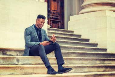Şık bir ceket, kravat, pantolon, kumaş ayakkabılar, merdivenlerde oturan, klasik ofis binasının önünde oturan, gülümseyen, cep telefonundaki mesajları kontrol eden genç bir iş adamı. Metin