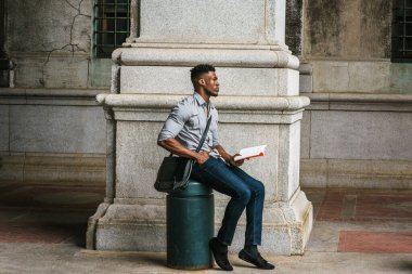Afrikalı Amerikan Koleji öğrencisi New York 'ta okuyor. Gri gömlek, kot pantolon, kumaş ayakkabılar, omuz derisi çanta, sokakta metal sütunda oturan siyah bir adam, kitap okuyor, düşünüyor.