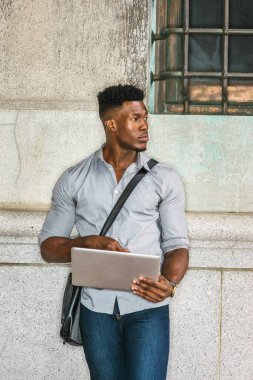 Afrikalı Amerikan Koleji öğrencisi New York 'ta okuyor. Gri gömlekli, kot pantolonlu, omuz derisi çanta taşıyan, sokakta duvara yaslanmış, okuyan, dizüstü bilgisayarla çalışan siyah bir adam.