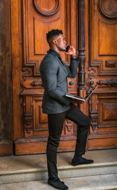 New York 'ta çalışan Afrikalı Amerikalı iş adamı. Modaya uygun ceketi, siyah kravatı, pantolonu, ayakkabıları, klasik tarz kapı aralığında duran, dizüstü bilgisayarla çalışan, telefonla arayan genç bir adam.
