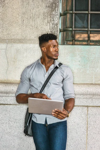 Afrikalı Amerikan Koleji öğrencisi New York 'ta okuyor. Gri gömlekli, kot pantolonlu, omuz derisi çanta taşıyan, sokakta duvara yaslanmış, okuyan, dizüstü bilgisayarla çalışan siyah bir adam.