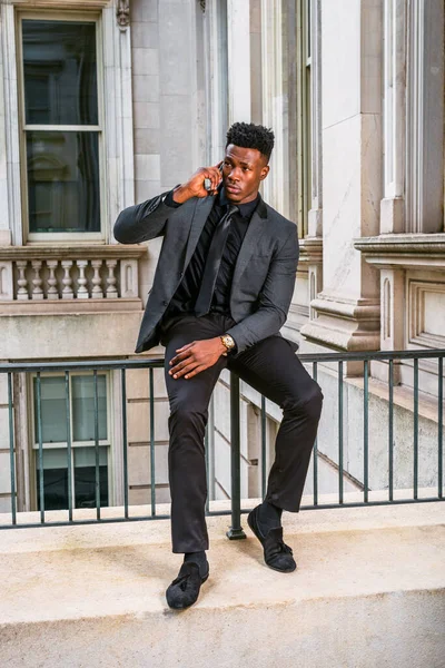 Şık bir ceket, siyah kravat, pantolon, kumaş ayakkabılar, kol saati takan, eski tarz ofis binasında korkuluklara oturan, cep telefonunu arayan genç bir iş adamı.