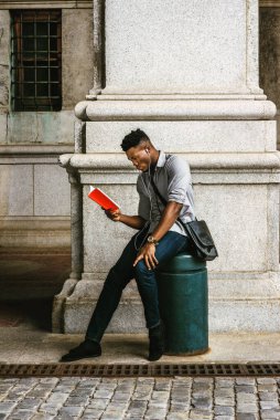 New York 'ta okuyan Afro-Amerikan Kolej öğrencisi, omuz çantası taşıyor, sokakta oturuyor, aşağı bakıyor, kırmızı kitap okuyor, kulaklıkla müzik dinliyor. filtrelenmiş etki