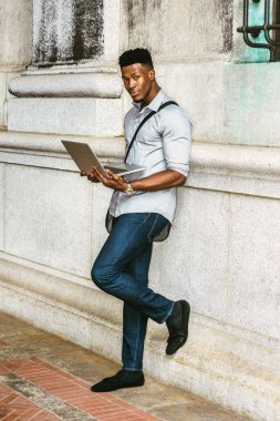 New York 'ta okuyan Afro-Amerikan Koleji öğrencisi, gömlek, kot pantolon giyen, omuz deri çanta taşıyan, kampüste duvarda duran, dizüstü bilgisayarla çalışan. filtrelenmiş etki