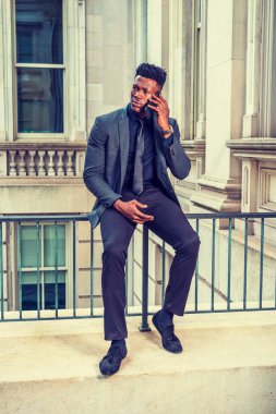 Şık bir ceket, siyah kravat, pantolon, kumaş ayakkabılar, kol saati takan, eski tarz ofis binasında korkuluklara oturan, cep telefonunu arayan genç bir iş adamı.