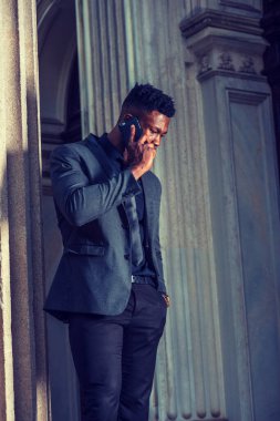 New York 'ta çalışan Afro-Amerikalı iş adamı, şık bir ceket, kravat takıyor, klasik ofis girişinin köşesinde duruyor, aşağıya bakıyor, cep telefonuyla konuşuyor. filtrelenmiş etki