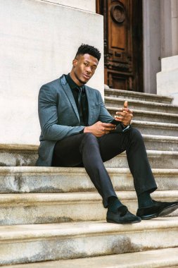 Afro-Amerikalı iş adamı seyahat ediyor, New York 'ta çalışıyor, şık bir ceket, kravat, pantolon, kumaş ayakkabılar giyiyor, klasik ofis binasının dışındaki merdivenlerde oturuyor, cep telefonuyla mesajlaşıyor..