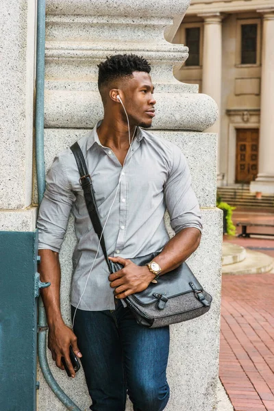 New York 'ta okuyan Afro-Amerikan üniversite öğrencisi, gri gömlek, kot pantolon, kol saati, omuz derisi çanta taşıyor, caddenin köşesinde duruyor, kulaklık ve cep telefonuyla müzik dinliyor.