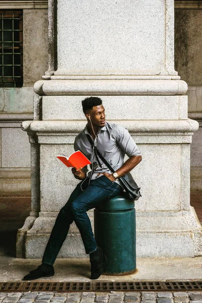 New York 'ta okuyan Afro-Amerikan Kolej öğrencisi, deri çanta taşıyor, sokakta oturuyor, kırmızı kitap okuyor, kulaklıkla müzik dinliyor, arkasına bakıyor. filtrelenmiş etki