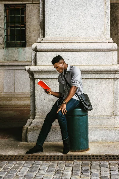 New York 'ta okuyan Afro-Amerikan Kolej öğrencisi, omuz çantası taşıyor, sokakta oturuyor, aşağı bakıyor, kırmızı kitap okuyor, kulaklıkla müzik dinliyor. filtrelenmiş etki