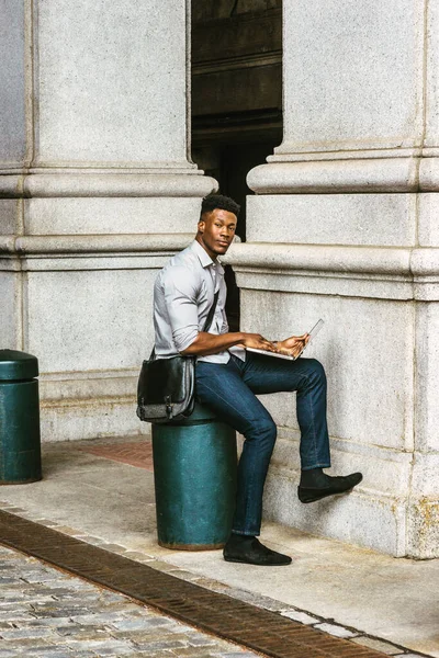 New York 'ta okuyan Afro-Amerikan Kolej öğrencisi, gömlek, kot pantolon giyiyor, omuz çantası taşıyor, sokakta bir sütunda oturuyor, okuyor, dizüstü bilgisayarla çalışıyor. filtrelenmiş effec