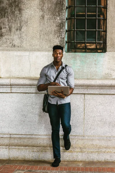 New York 'ta okuyan Afro-Amerikan Koleji öğrencisi, gömlek, kot pantolon giyen, omuz deri çanta taşıyan, kampüste duvarda duran, dizüstü bilgisayarla çalışan. filtrelenmiş etki