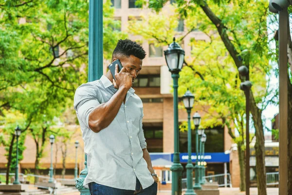 African American Man calling outside, wearing gray shirt, rolling over sleeves, standing against light pole on street in New York, lowering head, listening to cell phone. filtered effect