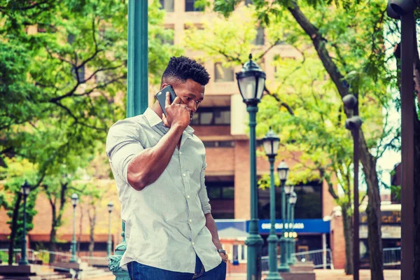 African American Man calling outside, wearing gray shirt, rolling over sleeves, standing against light pole on street in New York, lowering head, listening to cell phone.  filtered effect