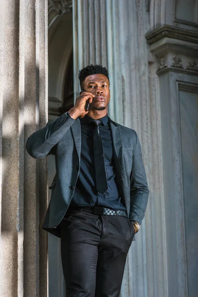 New York 'ta çalışan Afro-Amerikalı iş adamı, şık bir ceket giyiyor, siyah kravat takıyor, kol saati takıyor, klasik ofis girişinin köşesinde duruyor, cep telefonuyla konuşuyor. filtrelenmiş etki