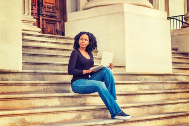 Afrikalı Amerikalı üniversite öğrencisi New York 'ta okuyor. Uzun kollu, V yakalı, kot pantolonlu, deri ayakkabılı, kampüsün dışındaki merdivenlerde oturan siyah bir kız, dizüstü bilgisayarla çalışıyor.