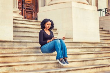 Afrikalı Amerikalı üniversite öğrencisi New York 'ta okuyor. Uzun kollu, V yakalı, kot pantolonlu, deri ayakkabılı, kampüsün dışındaki merdivenlerde oturan siyah bir kız, dizüstü bilgisayarla çalışıyor.