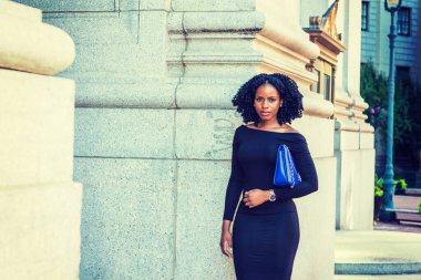 New York 'ta çalışan Afrikalı Amerikalı iş kadını. Uzun kollu, ince omuzlu, kolunun altında mavi çanta taşıyan, örgülü saç stili olan genç siyahi bir bayan, sana bakıyor.