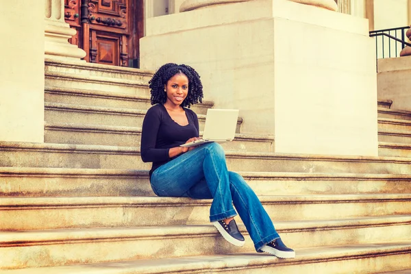 Afrikalı Amerikalı üniversite öğrencisi New York 'ta okuyor. Uzun kollu, V yakalı, kot pantolonlu, deri ayakkabılı, kampüsün dışındaki merdivenlerde oturan siyah bir kız, dizüstü bilgisayarla çalışıyor.