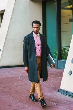 Sakallı, New York 'ta çalışan, yün paltolu, pembe gömlekli, kahverengi pantolonlu, deri çizmeli, papyonlu, dizüstü bilgisayar taşıyan,