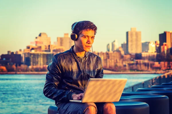 Asian American college student traveling, studying in New York, wearing black leather jacket, headphone, sitting by river, listening music, working on laptop computer. Far background is Brooklyn