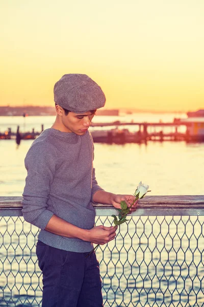 Asian American college student seeking love in New York, wearing newsboy cap, knitting sweater, holding white rose, standing by river in sunset, looking down, sad, thinking.