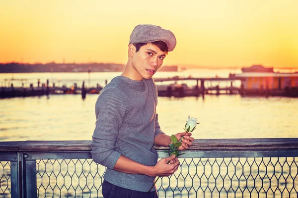 Asian American college student seeking love in New York, wearing newsboy cap, knitting sweater, holding white rose, standing by river in sunset, looking away, thinking. Filtered effect