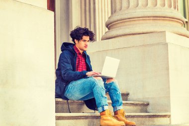 Çilli yüzlü, kıvırcık saçlı, kapüşonlu mavi ceketli, yırtık kot pantolonlu, sarı çizmeli, New York 'ta kampüste oturuyor, dizüstü bilgisayarla çalışıyor,.