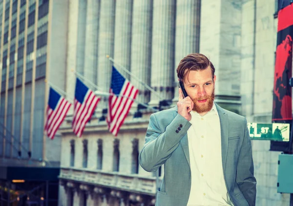 American Business Man with beard, mustache travels, works in New York. Guy wearing cadet blue suit, white shirt, stands on street with vintage buildings with flags, talks on cell phone.
