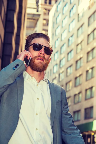 American businessman with beard, mustache traveling, working in New York, wearing cadet blue suit, white undershirt, sunglasses, walking through crowded high building street, talking on cell phone