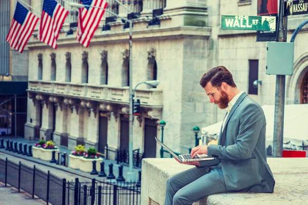 American Businessman with beard, mustache traveling, working in New York, wearing cadet blue suit, sitting on Wall Street, looking down, reading, working on laptop computer.
