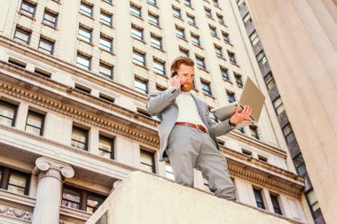 Sakallı, bıyıklı, New York 'ta çalışan, asker mavisi takım elbiseli, klasik binalarla sokakta duran, dizüstü bilgisayarda çalışan, cep telefonuyla konuşan Amerikalı iş adamı..