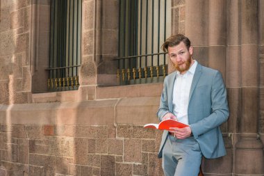 Sakallı, mavi takım elbiseli, beyaz tişörtlü, New York 'ta kampüste camları olan klasik duvarın karşısında duran, kırmızı kitap okuyan,.