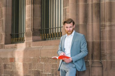 Sakallı, mavi takım elbiseli, beyaz tişörtlü, New York 'ta kampüste camları olan klasik duvarın karşısında duran, kırmızı kitap okuyan,.