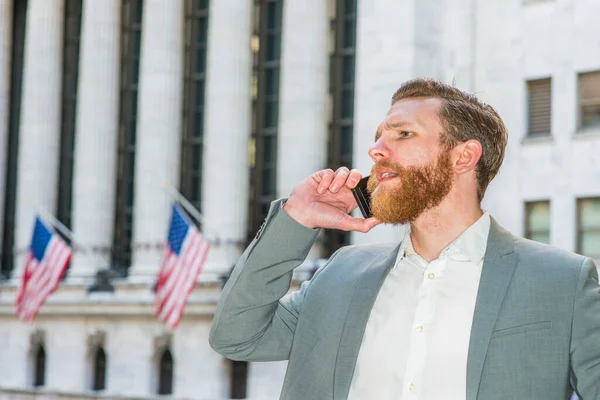 Young American Businessman with beard, mustache traveling, working in New York, wearing cadet blue suit, white shirt, standing on vintage street, talking on cell phone.