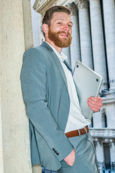 American Businessman with beard, mustache working in New York, wearing cadet blue suit, white undershirt, carrying laptop computer, standing against column outside vintage office building.