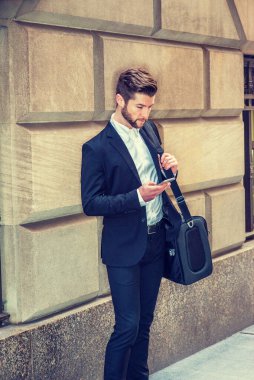 Sakallı, New York 'ta çalışan, siyah takım elbiseli, beyaz gömlekli, omuz çantalı, evrak çantalı, sokakta klasik duvarın yanında duran, cep telefonundan mesajları kontrol eden Amerikalı bir adam..