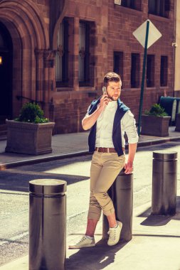 New York 'ta sıradan bahar modası. Sakallı, beyaz tişörtlü, kahverengi pantolonlu, spor ayakkabılı, mavi kazaklı, omuzlu, klasik binalı bir sokakta oturan, cep telefonuyla konuşan bir adam..