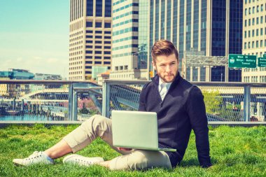 New York 'ta okuyan Amerikalı bir öğrenci seyahat ediyor. Siyah kazak, kahverengi pantolon, beyaz spor ayakkabı, sakallı bir adam, iş bölgesinde yeşil çimlerde oturuyor, dizüstü bilgisayarla çalışıyor..