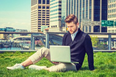 New York 'ta okuyan Amerikalı bir öğrenci seyahat ediyor. Siyah kazak, kahverengi pantolon, beyaz spor ayakkabı, sakallı bir adam, iş bölgesinde yeşil çimlerde oturuyor, dizüstü bilgisayarla çalışıyor..