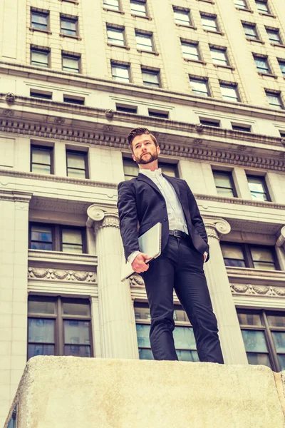 Businessman with beard working in New York, wearing black suit, white shirt, holding laptop computer, standing outside vintage office building, confident, success.
