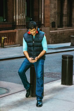 Modern Şehir Hayatı. Doğu Hindistan Amerikan Koleji öğrencisi uzun kollu tişört giyiyor, mavi yelek, kot pantolon, deri ayakkabı giyiyor, New York 'ta sokak sütununda oturuyor, mesajlaşıyor.. 
