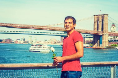 Doğu Hint asıllı, V yaka tişörtlü, kot pantolonlu, elinde beyaz bir gül, gülümsüyor ve seni bekliyor. Manhattan, Brooklyn köprüleri, arka planda tekne. 