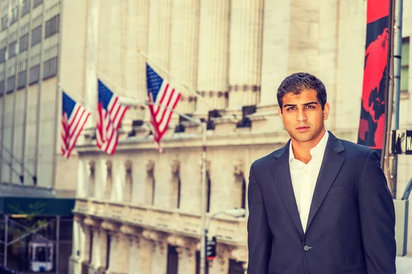 East Indian American Business Man traveling, working in New York. Wearing black suit, a college student standing on street with vintage buildings with flags, looking at you.