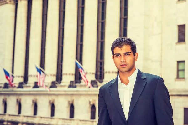 East Indian American Business Man traveling, working in New York. Wearing black suit, a college student standing on street with vintage buildings with flags, looking at you.