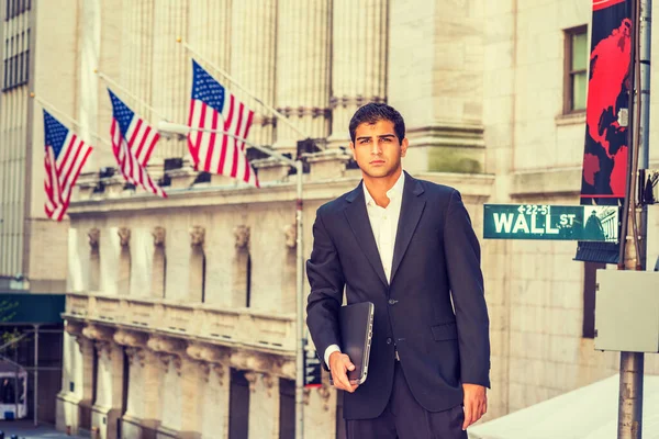 East Indian American Business Man traveling, working in New York. Wearing black suit, holding laptop computer, a student standing on Wall Street outside office building.