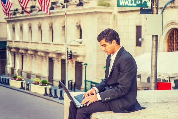 East Indian American Business Man traveling, working in New York. Wearing black suit, a college student sitting on Wall Street outside office, working on laptop computer.