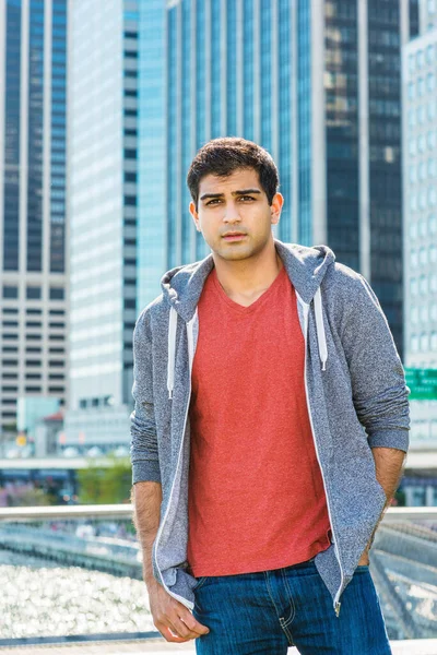 Portrait of City Boy. East Indian American college student traveling in New York, wearing red v neck T shirt, gray full zip hooded sweatshirt, jeans, standing in business district with high buildings.