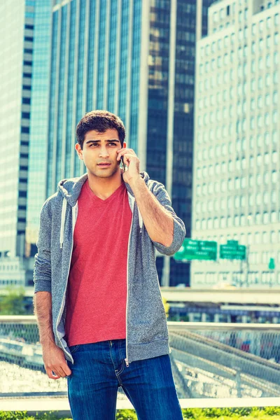 East Indian American college student traveling in New York, wearing red v neck shirt, gray hooded sweatshirt, standing in front of business district, talking on cell phone.