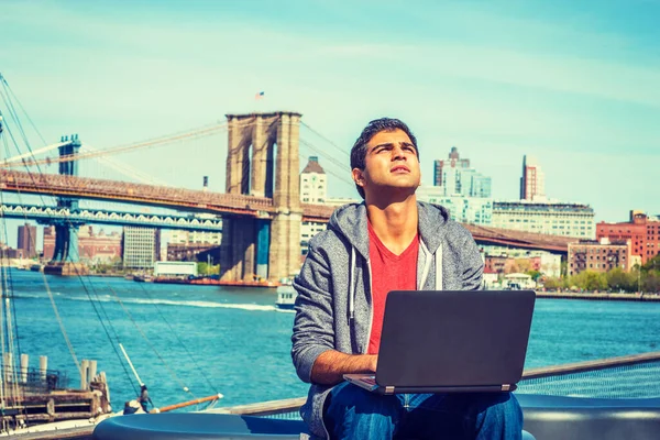 East Indian American college student traveling, studying in New York, wearing hooded sweatshirt, sitting by river, working on laptop computer, looking up, thinking hard. Brooklyn bridge on background